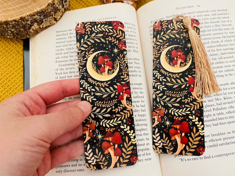 May include: A black bookmark with a gold crescent moon and mushrooms. The bookmark has a gold tassel.