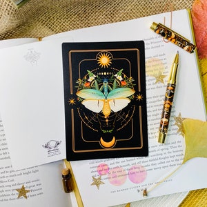 May include: A black card with a gold border features a detailed illustration of a moth with a green and blue body and white wings. The moth is surrounded by a circle of gold stars and a crescent moon. The card also features a sun and a circle of smaller moons.