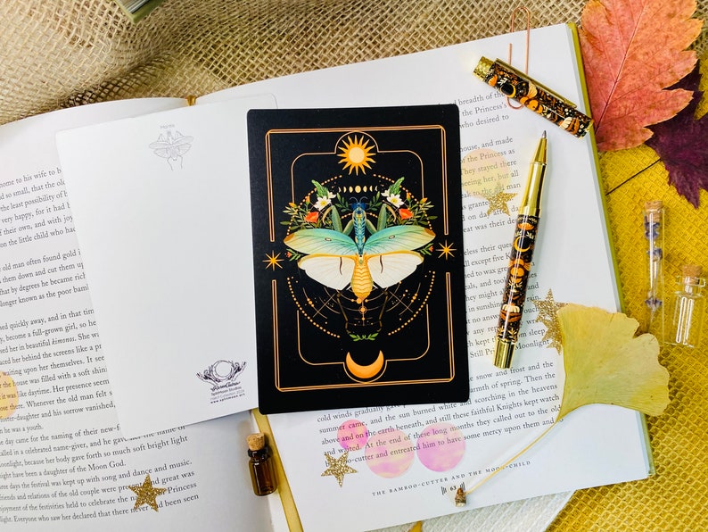 May include: A black card with a gold border features a detailed illustration of a moth with a green and blue body and white wings. The moth is surrounded by a circle of gold stars and a crescent moon. The card also features a sun and a circle of smaller moons.