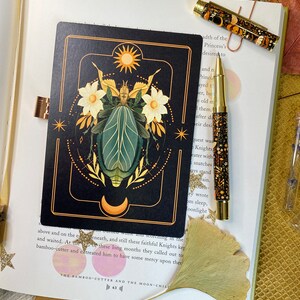 Peut inclure: Une carte de tarot noire avec des accents dor&eacute;s repr&eacute;sentant une illustration stylis&eacute;e d'un scarab&eacute;e avec des d&eacute;tails floraux et un soleil et une lune en arri&egrave;re-plan. Le texte de la carte est "The Bamboo-Cutter and the Moon-Child".