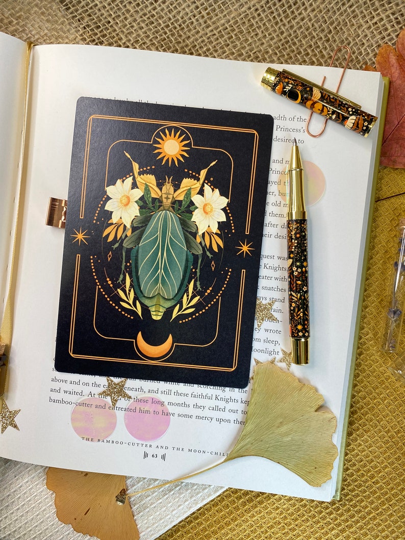 Peut inclure: Une carte de tarot noire avec des accents dor&eacute;s repr&eacute;sentant une illustration stylis&eacute;e d'un scarab&eacute;e avec des d&eacute;tails floraux et un soleil et une lune en arri&egrave;re-plan. Le texte de la carte est "The Bamboo-Cutter and the Moon-Child".