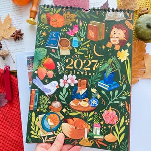 May include: A 2027 wall calendar with a dark green background featuring colorful illustrations of a hedgehog, a fox, books, coffee cups, strawberries, flowers, and a person reading. The calendar has a spiral binding at the top and gold text.