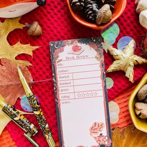 K&ouml;nnte beinhalten: Ein Notizblock f&uuml;r Buchrezensionen mit dekorativem Rand, mit Feldern f&uuml;r Titel, Autor und Bewertung. Ein goldener und schwarzer Stift mit floralem Design liegt neben dem Notizblock. Der Hintergrund umfasst Herbstbl&auml;tter, Eicheln und dekorative Schalen.