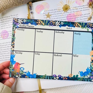 Op de afbeelding: Een weekplanner met een bloemenontwerp. De planner heeft secties voor maandag, dinsdag, woensdag, donderdag, vrijdag, weekend, prioriteit en notities. De planner is versierd met een kolibrie en bloemen.