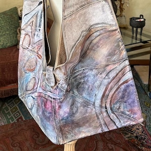 May include: A large, shoulder bag made of distressed leather. The bag features a unique, abstract design with shades of purple, blue, and pink. The bag is displayed on a mannequin, showcasing its shape and size. The bag's strap is wide and appears comfortable.
