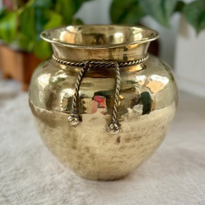 Könnte beinhalten: Eine gehämmerte Messingvase mit einer Kordelverzierung und dekorativen Kugelakzenten. Die Vase hat einen runden Körper und eine breite Öffnung.