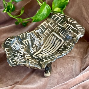 May include: A black and white ceramic dish with a geometric design. The dish has a unique shape and a single leg.