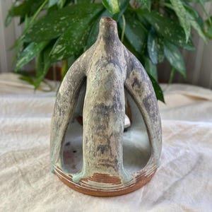 May include: A handmade ceramic sculpture with a unique, abstract design. The piece features a light green and gray color palette with a textured surface. The base is a reddish-brown color. The sculpture has an open, airy design.