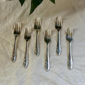 May include: Seven silver forks displayed on a cream-coloured cloth. The forks feature a simple, elegant design with a decorative element at the base of the handle. The tines are evenly spaced.