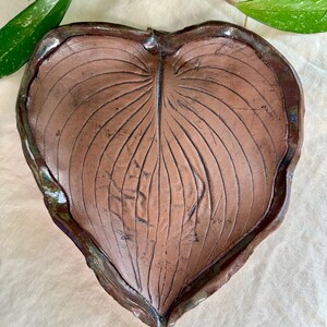 May include: A handcrafted, heart-shaped ceramic leaf dish. The brown dish features detailed vein patterns and a dark, glossy rim. The leaf dish is a decorative piece, perfect for holding small items or as a unique home accent.