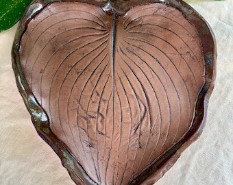 May include: A handcrafted, heart-shaped ceramic leaf dish. The brown dish features detailed vein patterns and a dark, glossy rim. The leaf dish is a decorative piece, perfect for holding small items or as a unique home accent.