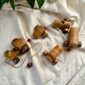 May include: A set of four wooden toy train cars in various shapes and sizes, connected by a string. The cars are primarily light brown with darker brown wheels and accents. Small red beads are interspersed along the string.