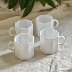 Peut inclure: Quatre tasses blanches de forme géométrique avec des anses arrondies sont disposées sur une surface texturée de couleur crème. Les tasses ont une finition mate et un design moderne. Du feuillage vert est visible en arrière-plan.