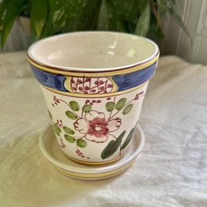 Peut inclure: Un pot de fleurs en céramique couleur crème avec sa soucoupe assortie. Le pot présente un motif floral peint à la main dans des tons de rose, vert et rouge, avec une large bande bleue sur le bord rehaussée de lignes jaunes et d'un quadrillage.