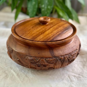 May include: A round wooden container with a lid. The container is a warm brown colour and has carved details around the base. The lid has a small knob. The container is likely used for storage or display.