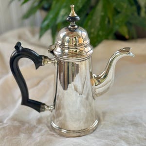 Puede incluir: Una cafetera plateada con un asa negra y una parte superior decorativa. La cafetera tiene un pico curvo y se asienta sobre una superficie blanca. El asa es curva y la parte superior tiene un acento dorado y negro.