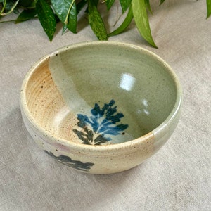 Puede incluir: Un pequeño cuenco de cerámica blanca con un diseño floral azul y marrón. El cuenco tiene un esmalte moteado y un borde ligeramente irregular.