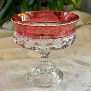 Puede incluir: Un cuenco de pedestal de vidrio transparente con un borde rojo. El cuenco tiene un diseño decorativo y texturizado en la base y el borde. El cuenco es probablemente vintage y adecuado para servir postres o exhibir objetos pequeños.