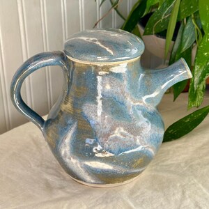 May include: A handmade ceramic teapot with a lid. The teapot is glazed in shades of blue, white, and gold, creating a marbled effect. It features a curved handle and a spout for pouring. The teapot is a decorative item.