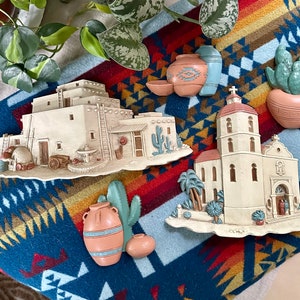 Könnte beinhalten: Ein Set aus fünf Keramik-Wanddekorationen mit Südwest-Thema. Das Set beinhaltet eine Kirche, einen Kaktus, ein Gebäude mit einer Leiter, einen Topf mit einem Kaktus und einen Topf mit einer Blume. Die Teile sind in Weiß-, Braun- und Grüntönen bemalt.