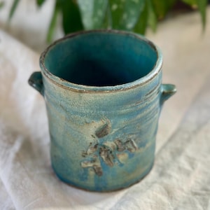 May include: A handmade ceramic teal-colored pot with two small handles. The pot has a textured surface with a floral design. The interior is a darker shade of teal. The pot is suitable for holding small plants or as a decorative item.