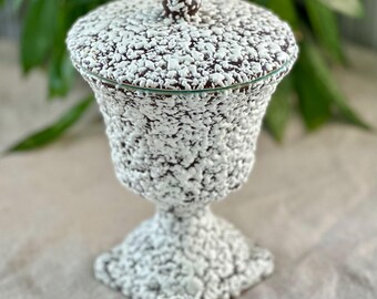 May include: A white ceramic jar with a lid, featuring a textured, speckled design. The jar has a round lid with a knob on top and a wide, round base.