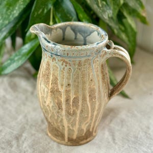 May include: A handmade ceramic pitcher with a spout and handle. The pitcher has a beige base with vertical streaks of white and blue glaze dripping down the sides. The rim is accented with a blue glaze.