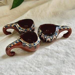 Może przedstawiać: Trzy ręcznie robione ceramiczne kubki z zakrzywionymi uchwytami. Kubki mają głęboki czerwonobrązowy kolor ze skomplikowanymi wzorami w kolorze niebieskim, kremowym i czarnym. Wnętrze kubków jest ciemniejsze.