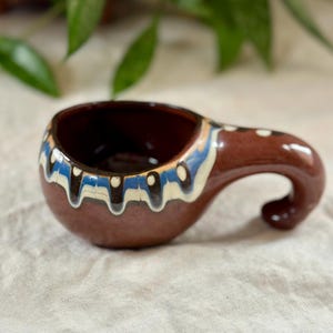 Puede incluir: Una taza pequeña de cerámica marrón con asa. La taza tiene un diseño pintado en blanco y azul con detalles negros.