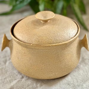 Könnte beinhalten: Eine beige Keramik-Auflaufform mit Deckel und zwei Griffen. Die Form hat eine strukturierte Oberfläche.