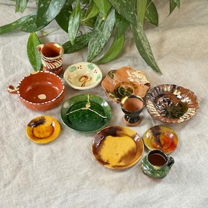 May include: A collection of miniature pottery dishes and cups in various colours and patterns, including green, brown, yellow, and orange. The dishes are arranged on a light brown fabric surface.