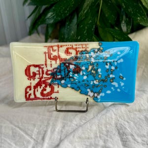 Könnte beinhalten: Rechteckige Glasschale mit cremefarbenem Grund und lebendigem Design. Die linke Seite zeigt rote Schrift, während die rechte Seite leuchtend blau mit weißen und goldenen Akzenten ist. Die Schale wird von einem kleinen Metallständer getragen.
