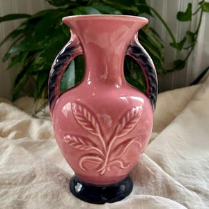 Könnte beinhalten: Eine rosa Keramikvase mit zwei Henkeln und einem dunkelblauen Sockel. Die Vase hat ein weißes Blattmuster und dunkelblaue Akzente an den Henkeln. Die Vase ist ein dekorativer Gegenstand.