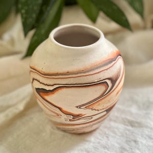 May include: A small, round ceramic vase with a cream-colored base and swirling patterns of brown and orange. The vase has a wide opening and a smooth, unglazed interior. The design resembles a marbled effect.