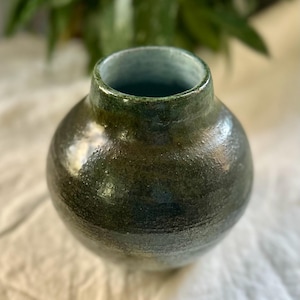 Puede incluir: Un jarrón de cerámica hecho a mano con un cuerpo redondeado y un cuello estrecho. El jarrón presenta un esmalte verde oscuro y marrón con sutiles variaciones de color. El interior tiene un tono verde más claro. Este jarrón decorativo es adecuado para la decoración del hogar.