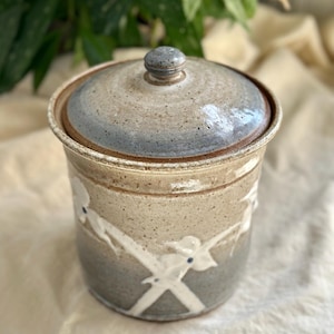 May include: A ceramic jar with a lid, featuring a speckled beige and blue glaze. White floral designs with blue accents adorn the jar's exterior. The jar is a cylindrical shape, suitable for kitchen or home decor.