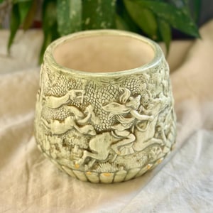 Puede incluir: Jardinera de cerámica verde claro con un relieve detallado de figuras y animales. La jardinera tiene una abertura ancha y una superficie texturizada. El diseño presenta figuras clásicas y animales en un bosque.