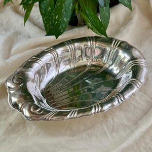 Puede incluir: Una fuente ovalada plateada con un diseño decorativo acanalado y un borde festoneado. La fuente tiene detalles ornamentados y un interior liso y reflectante. Se encuentra sobre un fondo neutro.