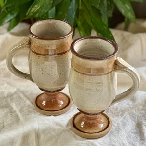 Puede incluir: Dos tazas de cerámica hechas a mano con bases de pedestal. Las tazas son de color beige con detalles y asas marrones. Los bordes y las bases tienen un esmalte marrón más oscuro. Las tazas se exhiben sobre una tela blanca, con follaje verde en el fondo.