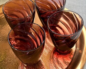 以下が含まれることがあります： 渦巻き模様の入った、透明で紫がかった小さな飲み物が4つ、装飾的なデザインの金色のトレイの上に置かれています。