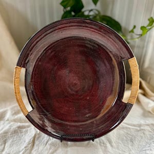 May include: A burgundy ceramic serving platter with woven handles. The round platter has a glossy finish and is supported by a black metal stand. The handles are made of a light brown woven material.