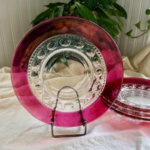 May include: A set of vintage glass plates with a vibrant magenta rim and a clear, textured center. The plates are displayed on a metal stand, showcasing their design. The plates have a decorative pattern around the center.