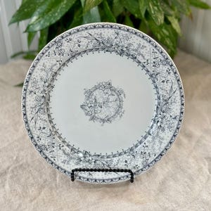 May include: A white ceramic plate with a black and grey floral pattern. The plate has a scalloped edge and a raised centre design.