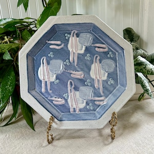 Puede incluir: Plato octogonal de cerámica con esmalte azul y blanco. El plato presenta un diseño geométrico con acentos rosados. El plato está sobre un soporte de metal dorado.