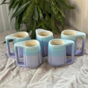 May include: Set of five ceramic mugs with a gradient design in shades of blue and purple. The mugs have a unique handle design.