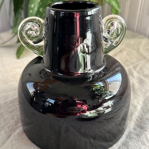 Może przedstawiać: Czarny szklany wazon z dwoma przezroczystymi szklanymi uchwytami. Wazon ma zaokrąglone ciało i szeroki otwór.