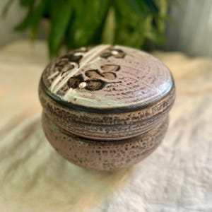 Könnte beinhalten: Eine handgefertigte Keramik-Schmuckdose mit Deckel. Die Dose hat eine gedämpfte rosa Farbe mit einer strukturierten Oberfläche. Der Deckel hat ein Blumendesign in Braun und Weiß. Die Dose hat einen Durchmesser von ca. 10 cm und ist 8 cm hoch.