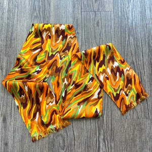 May include: A colorful scarf with a wavy pattern in shades of orange, brown, yellow, and green. The scarf has a fringed edge and is made of a silky material. The scarf is a fashion accessory.