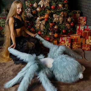 May include: A large, plush, pale blue spider toy with long, furry legs and a white underside. The toy is on a brown rug, with a Christmas tree and wrapped gifts in the background. The spider toy is a unique decorative item.