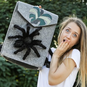 May include: Grey felt backpack with a large, black, plush spider applique. The flap features a leaf design in shades of green and grey. The bag has a black strap and a black base.